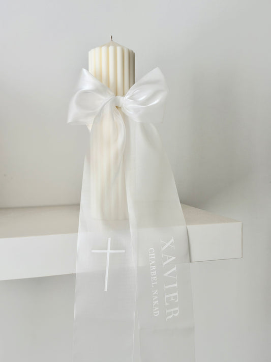 A cream-colored ribbed pillar candle tied with a sheer white organza ribbon bow. The ribbon is printed with a white cross and the text 'XAVIER CHARRLES NAKED'.