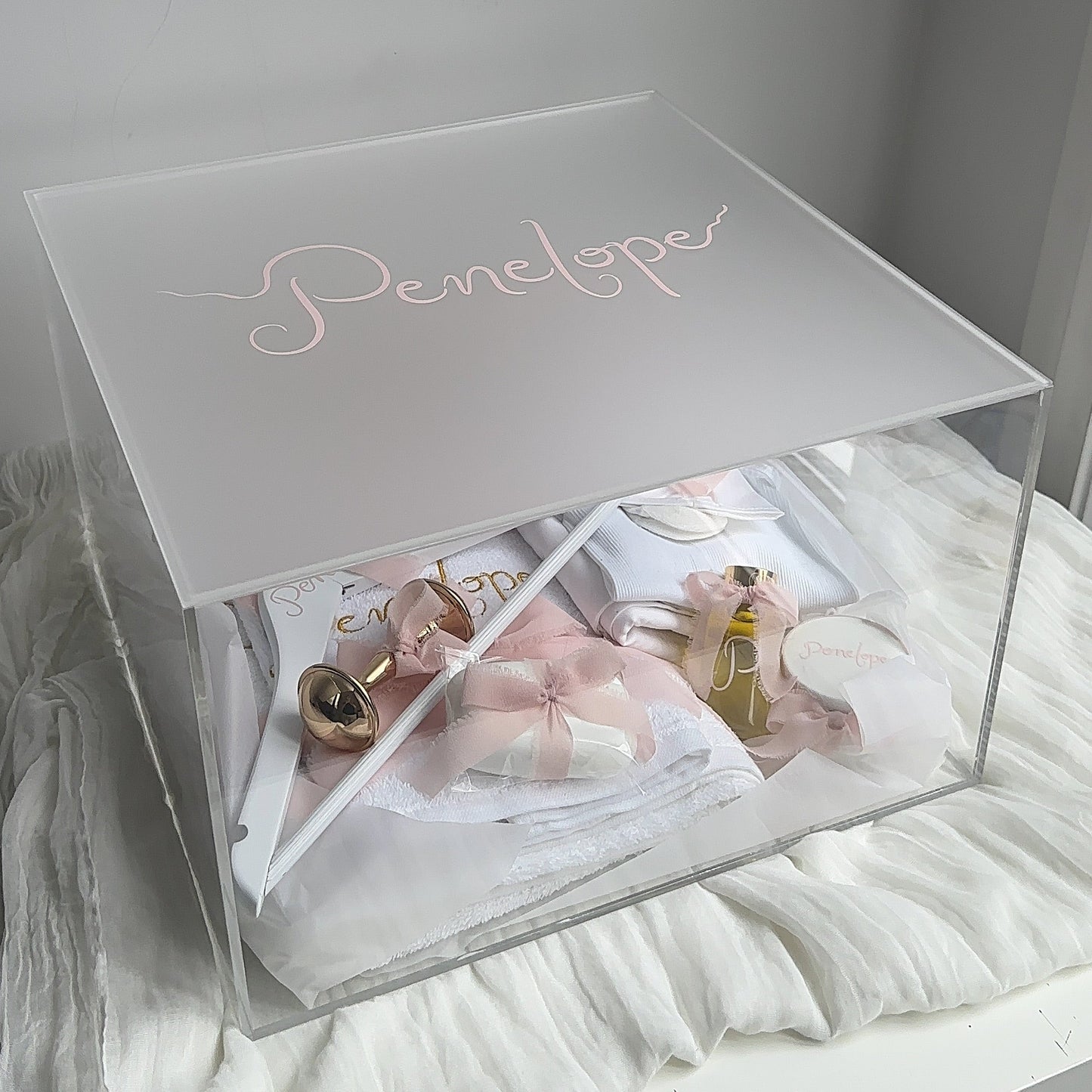 A clear acrylic jewelry christening box with a frosted lid engraved with the name 'Penelope' in rose gold script. The box is shown open, filled with white and pink jewelry boxes and small accessories, resting on a white fabric-covered surface against a light gray wall.