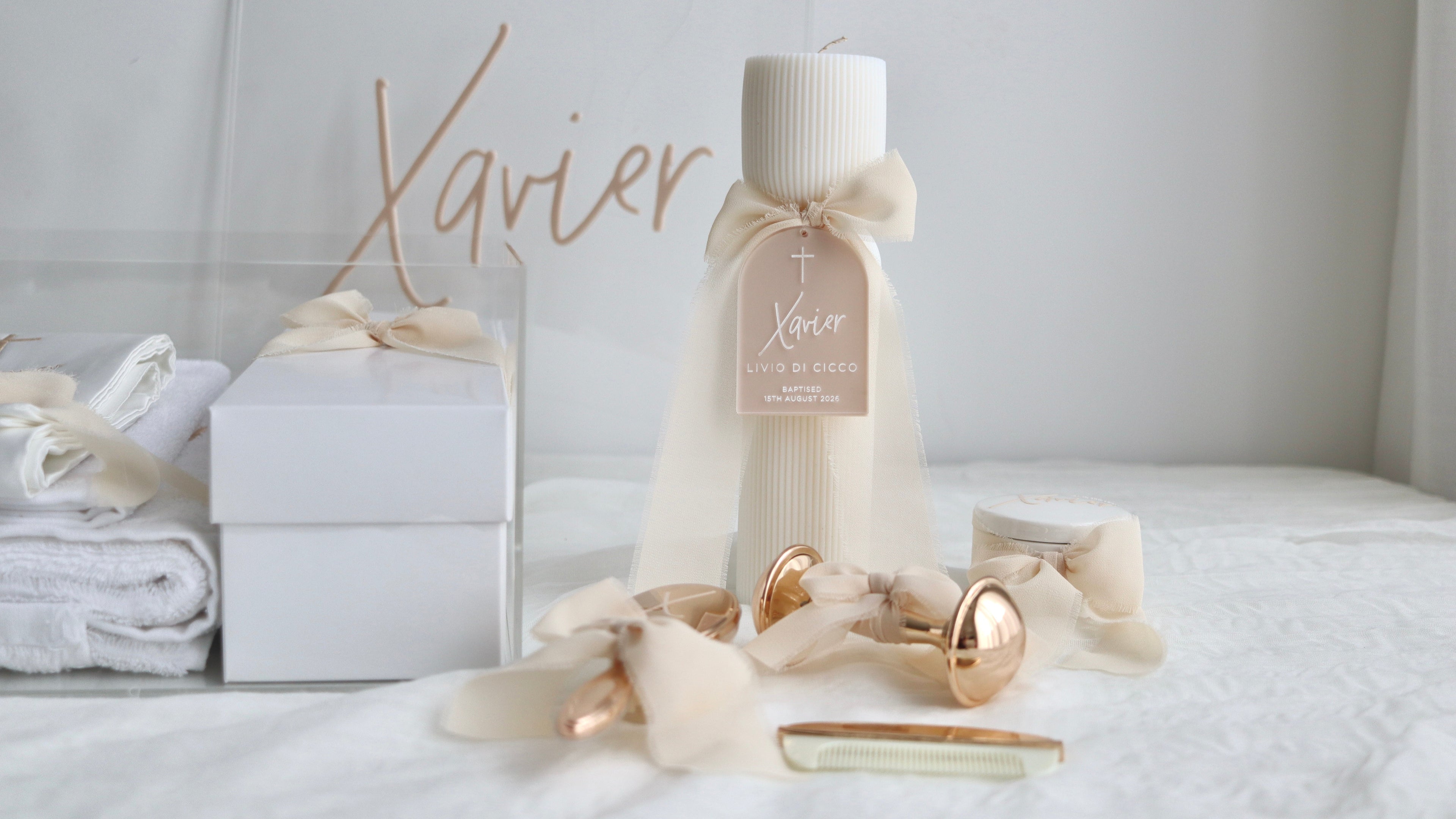 A personalized christening gift set featuring a ribbed white pillar candle with a cream bow and tag, a white gift box, folded white towels, and gold rattle and ribbon accents on a white bed.