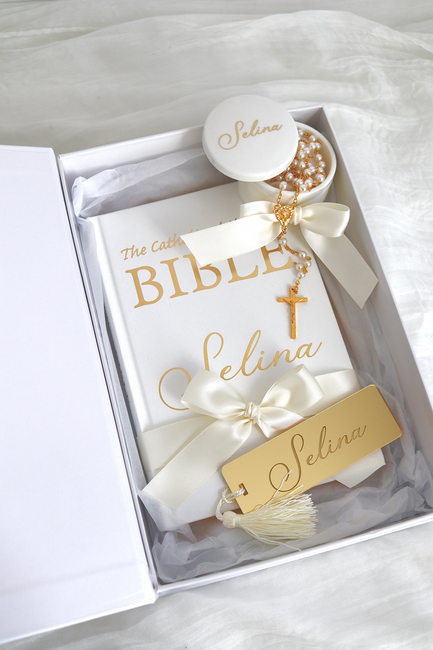 A personalized Catholic Bible gift set in a white box, featuring a white Bible with gold lettering, cream satin ribbons, a gold cross, a pearl rosary, a small white candle, and a gold tag all personalized with the name 'Selina'.