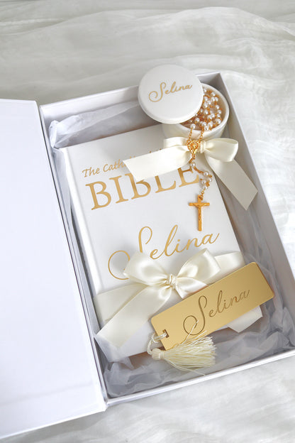 A personalized Catholic Bible gift set in a white box, featuring a white Bible with gold lettering, cream satin ribbons, a gold cross, a pearl rosary, a small white candle, and a gold tag all personalized with the name 'Selina'.
