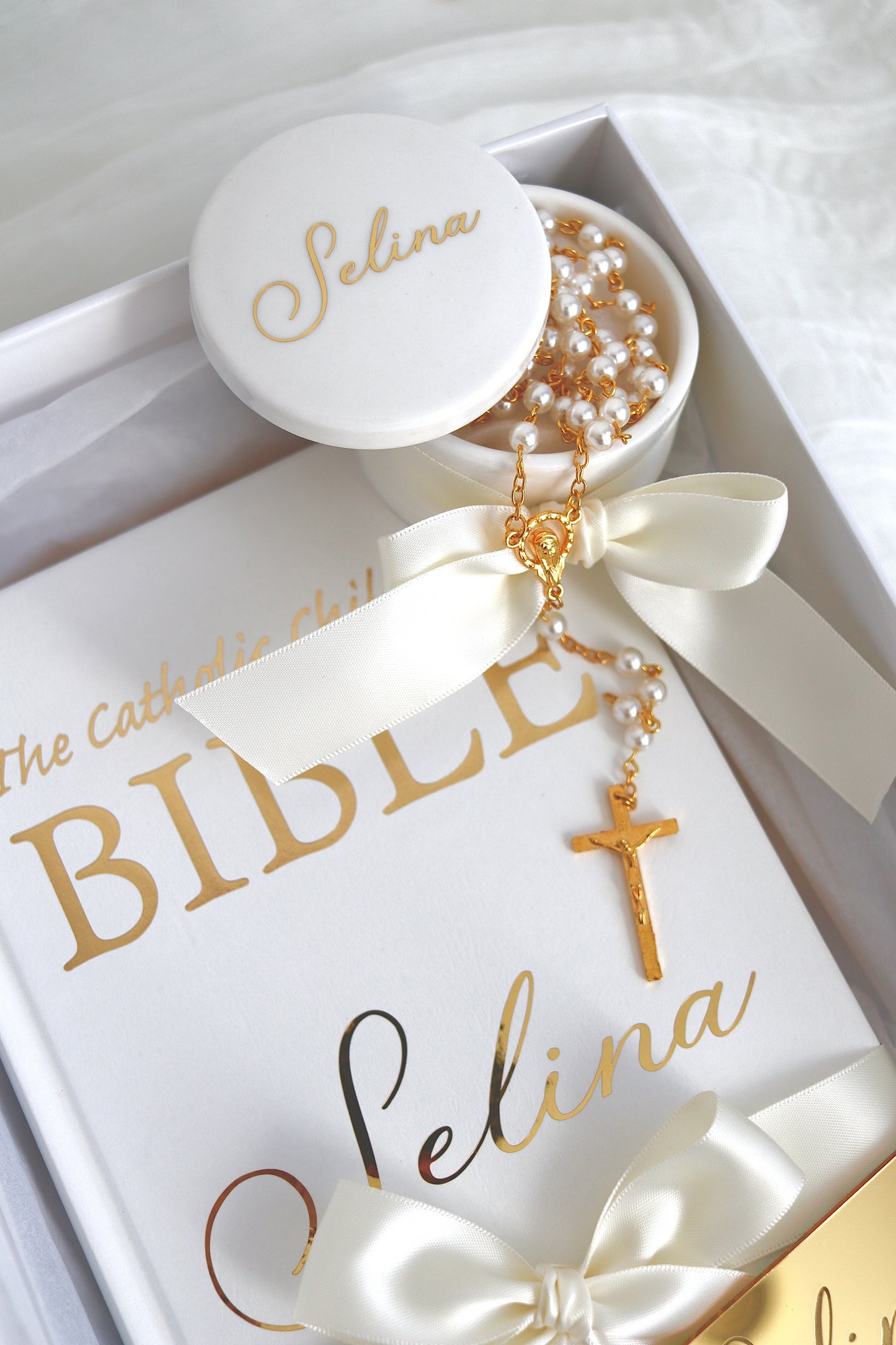 A close-up of a gift set featuring a gold and pearl rosary with a cross and nameplate engraved 'Selina', displayed on top of a white Bible with gold lettering and matching white and gold ribbons.