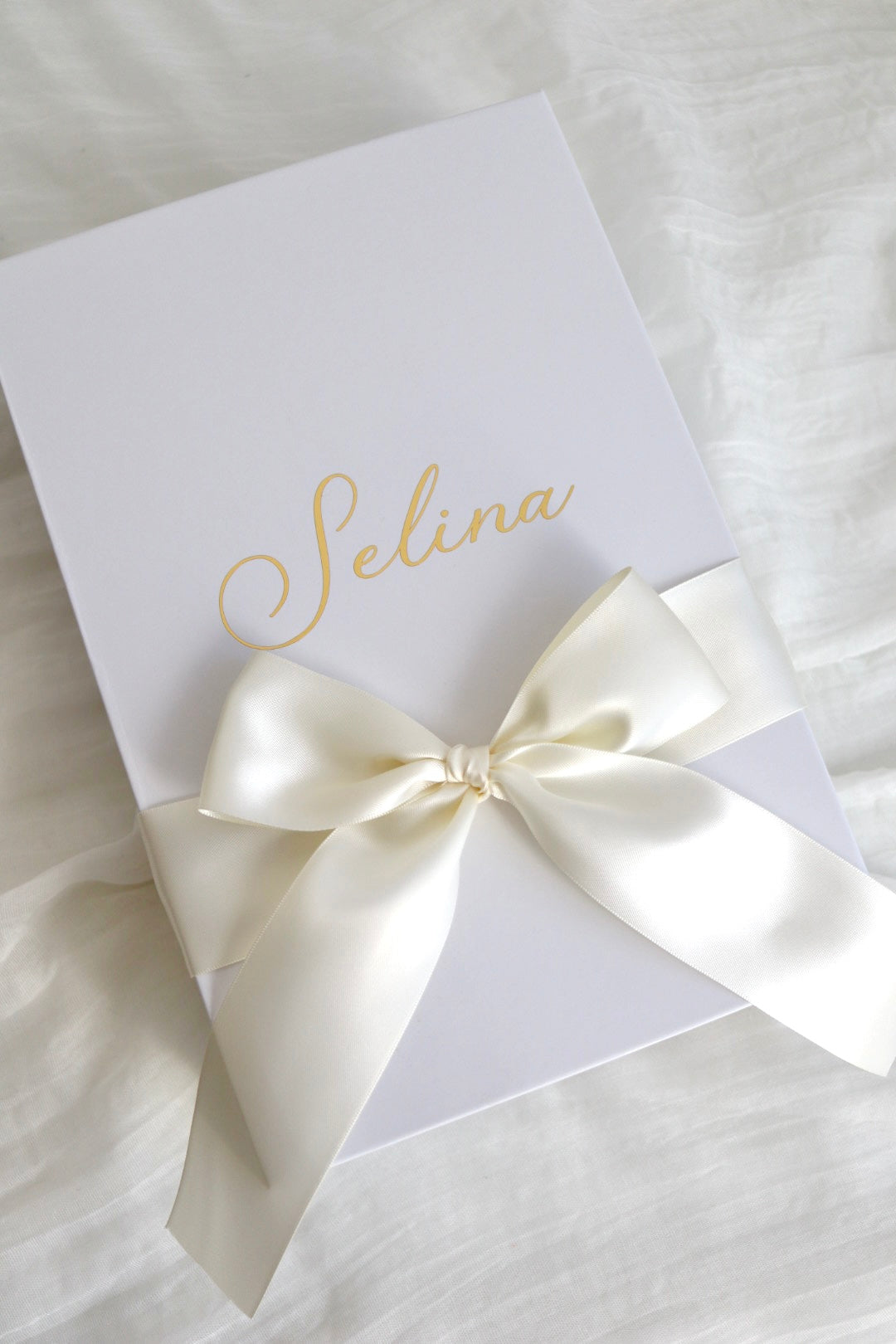 A white rectangular gift box or package tied with a large cream satin ribbon bow, placed on a white fabric surface. The box is printed with the name 'Selina' in elegant gold script.