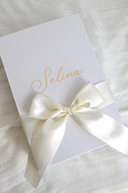 A white rectangular gift box or package tied with a large cream satin ribbon bow, placed on a white fabric surface. The box is printed with the name 'Selina' in elegant gold script.