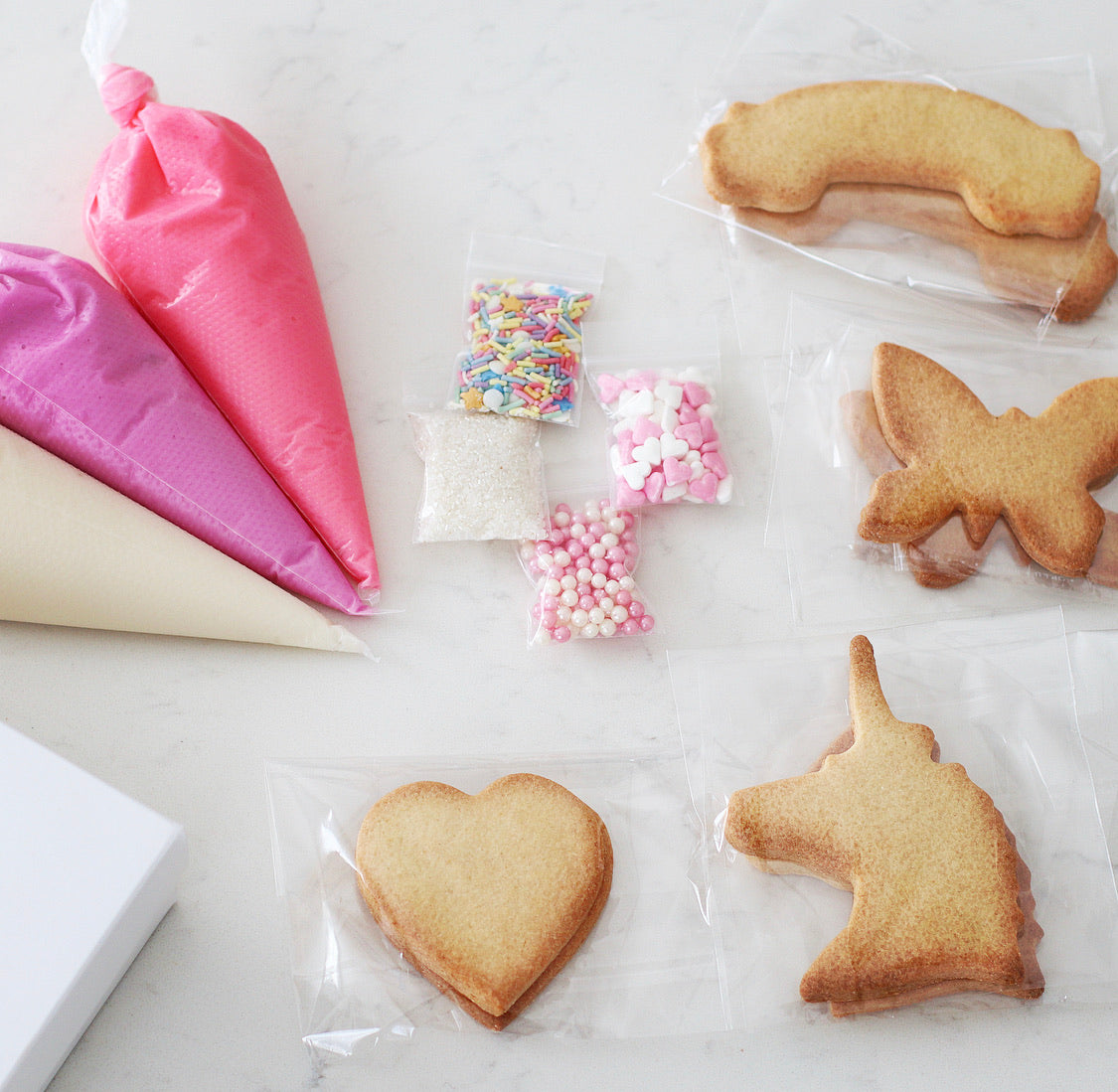 Unicorn & Rainbows DIY Cookie Kit - Contains 3 Piping bags, filled with ready to use icing in Pink, Purple & White.  4 Bags of sprinkles/decorations  8 Cookies, ready to decorate. Cookie shapes include: Rainbow, Butterfly, Unicorn & Heart.